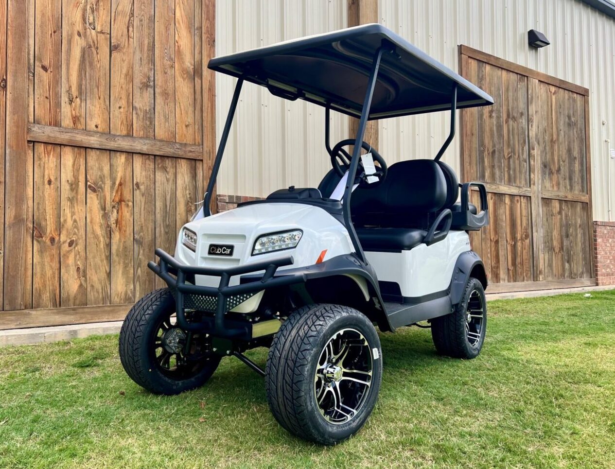White golf cart with black roof parked on grass near wooden wall.