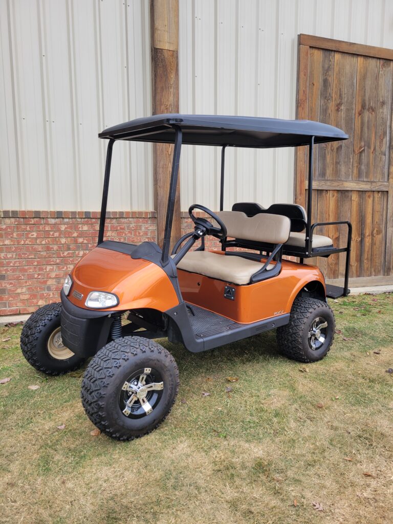 Orange golf cart parked on grass near a wooden fence.