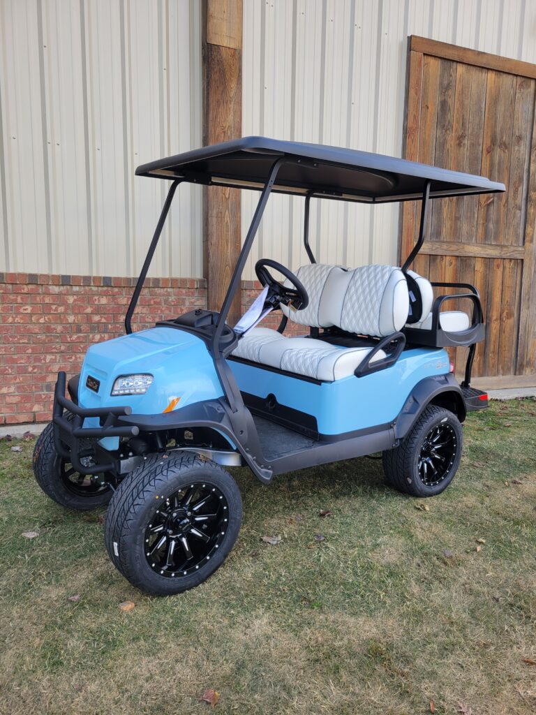 Blue golf cart with black roof and custom wheels parked on grass.