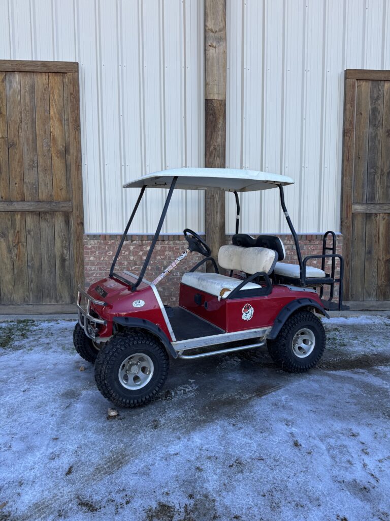 Red golf cart parked outside on snowy ground near wooden doors.