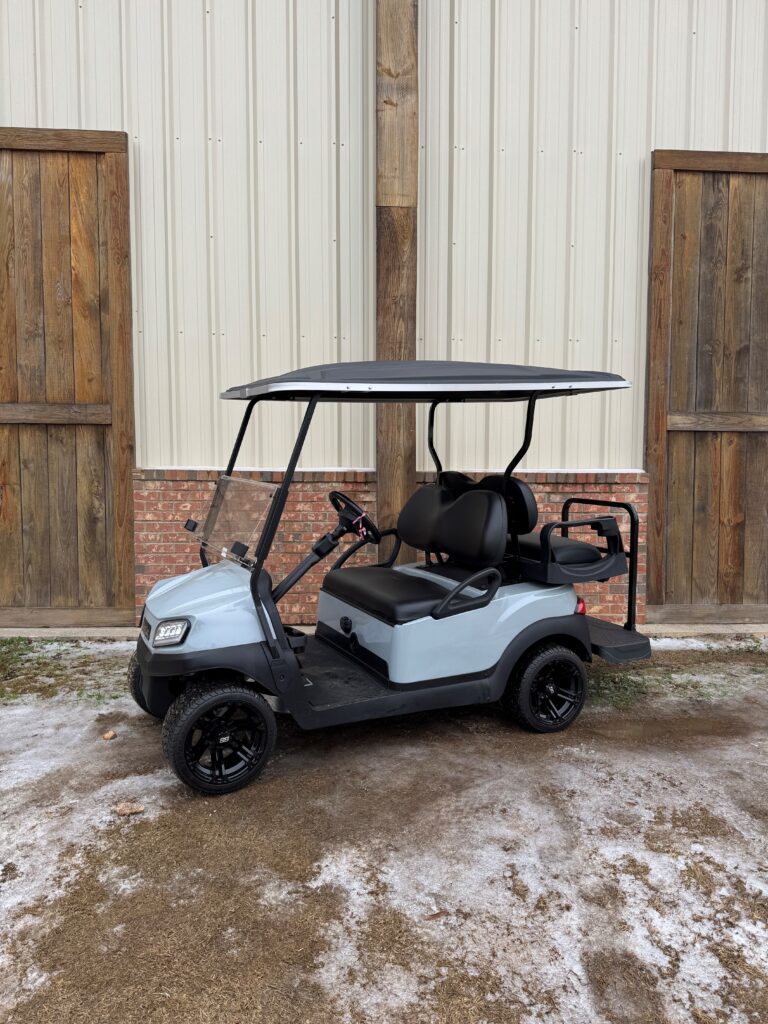 A gray golf cart parked on a snowy surface near wooden doors.
