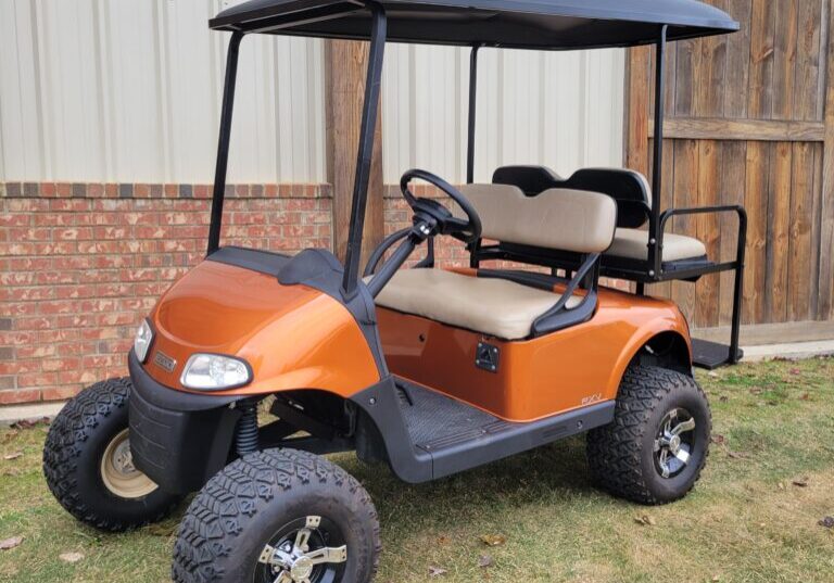 Orange golf cart parked on grass near a wooden fence.