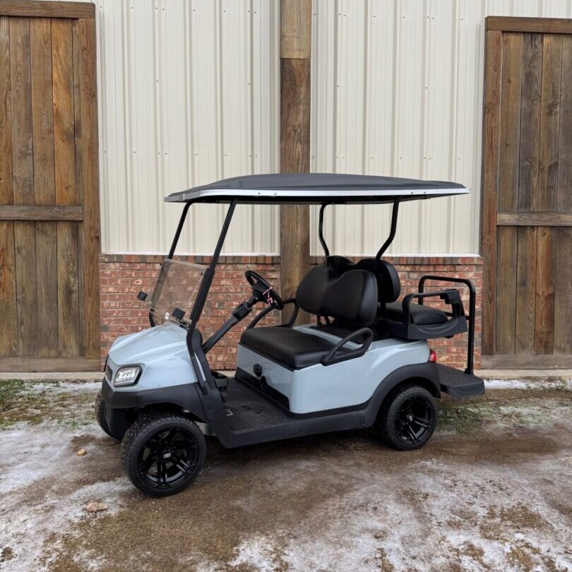 A gray golf cart parked on a snowy surface near wooden doors.
